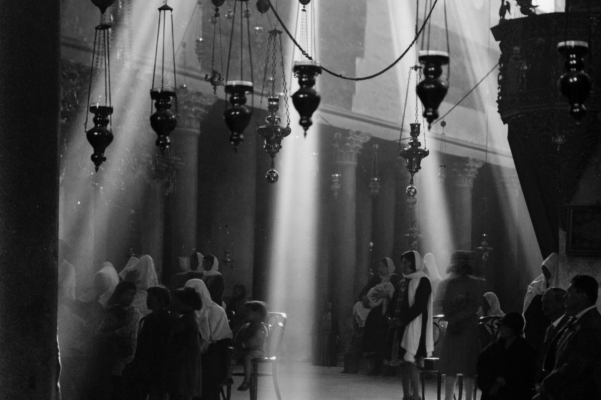 Pilgrims inside the Basilica of the Nativity in Bethlehem, sunlight streaming past hanging lamps and columns