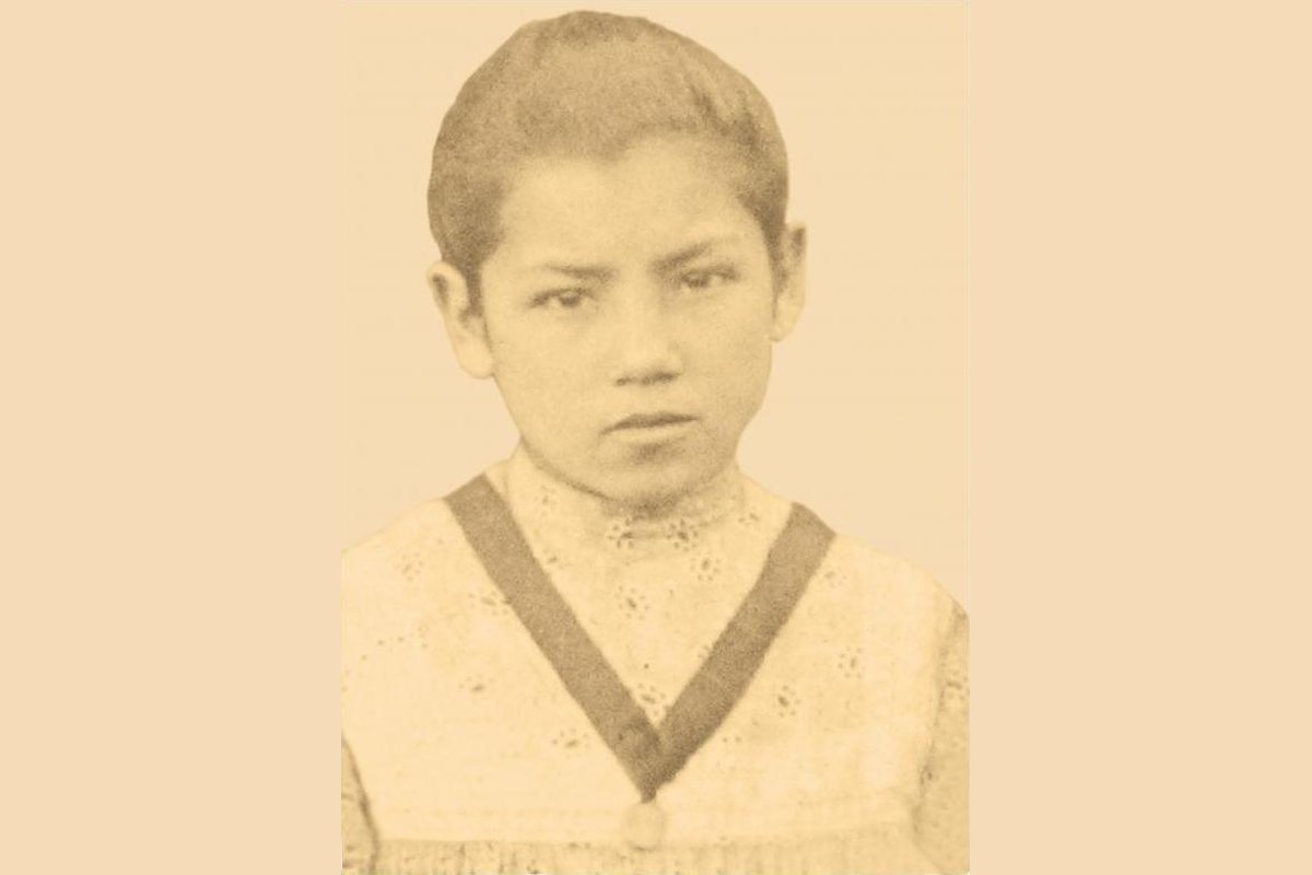 A sepia-toned period photograph of Laura Vicuña as a young girl, wearing a light pinafore-style dress with a dark sash, looking directly at the camera.