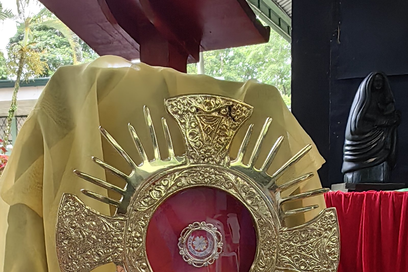 A gold reliquary monstrance holding a small relic of Saint Carlo Acutis in its central medallion, displayed on a yellow cloth during a public veneration