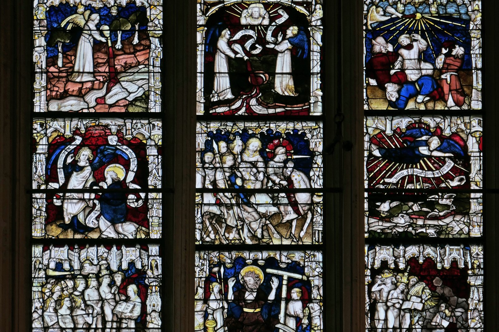 A large Gothic cathedral stained glass window divided into many small colored panels, each depicting a separate biblical scene, glowing with reds, blues, and golds.