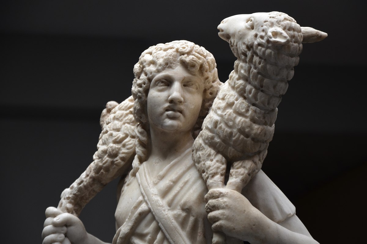 Eine verwitterte Marmorstatue eines jungen Mannes, der ein Lamm über seinen Schultern trägt und dessen Beine mit beiden Händen hält.