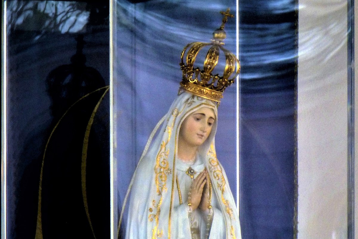 Eine Nahaufnahme einer Statue der Jungfrau Maria mit goldener Krone und weißen, goldverzierten Gewändern, die Hände im Gebet gefaltet.