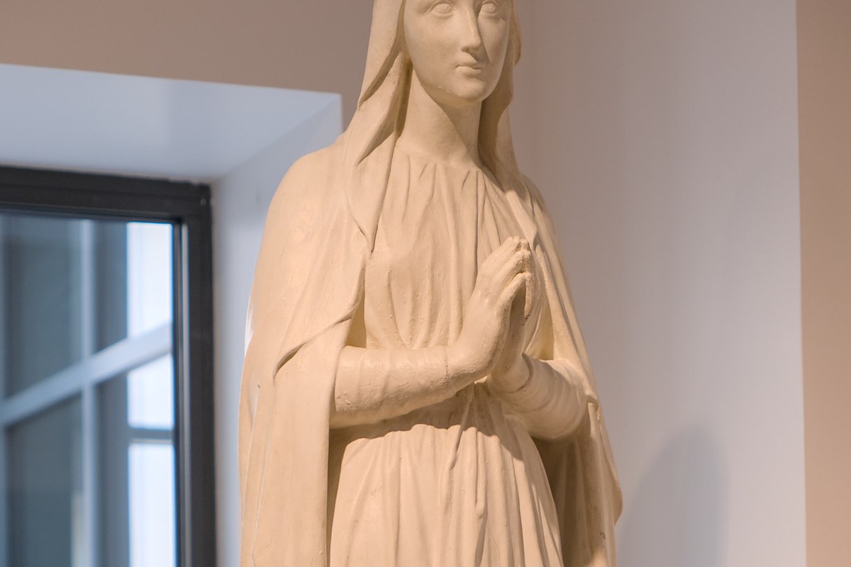 A close-up of a serene white plaster statue of a woman with hands folded in prayer, wearing a veil.
