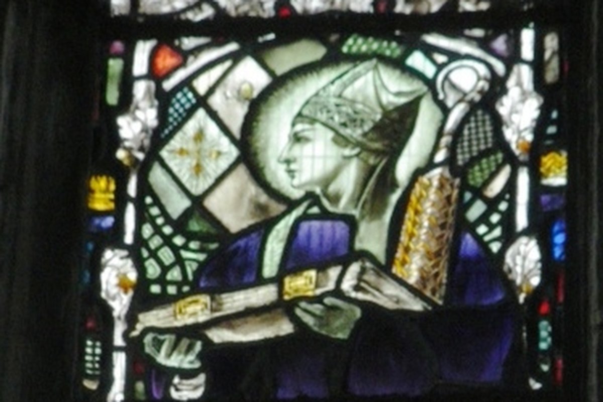 A stained-glass window showing a bishop saint in profile, wearing a mitre and holding an open book, with the name Aidan inscribed on a banner below.
