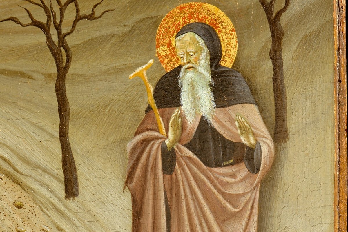 A tempera panel painting of a haloed, white-bearded hermit monk in a rose-and-black habit standing in a bare, wintry landscape with dead trees and grazing deer, a small monastery visible in the distance.