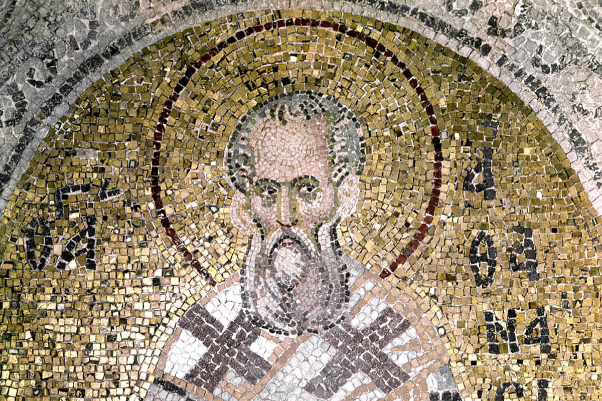 A Byzantine mosaic icon of a bearded bishop saint with a gold halo, wearing a cross-patterned omophorion and holding a Gospel book.