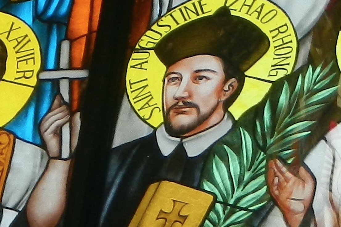 A stained-glass window portrait of Saint Augustine Zhao Rong, shown in traditional Chinese dress holding a Bible and a martyr's palm branch.