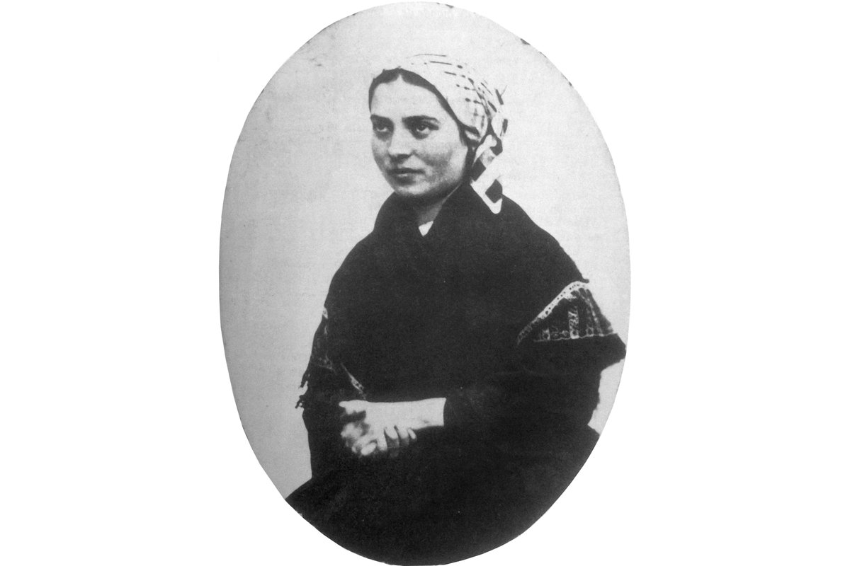 A black-and-white oval studio photograph of a young Bernadette Soubirous, wearing a patterned headscarf and a dark shawl, hands folded, looking calmly to one side.