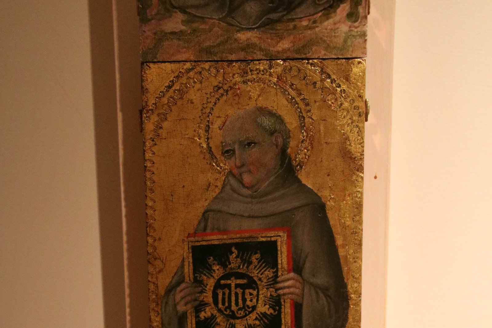 A gold-ground panel painting of Saint Bernardine of Siena in Franciscan grey habit, holding his signature wooden tablet bearing the radiant IHS monogram of the Holy Name of Jesus.