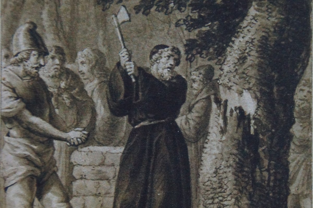 An 18th-century engraving of a robed missionary swinging an axe into a great oak tree while armed men and onlookers watch.
