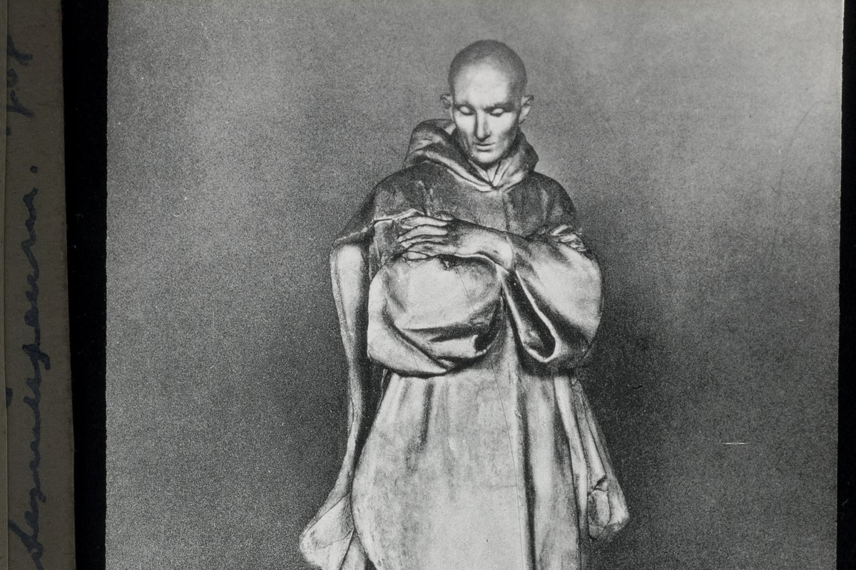 A photograph of a sculpted model of a bald, robed monk standing with arms crossed over his chest and head bowed in contemplation.