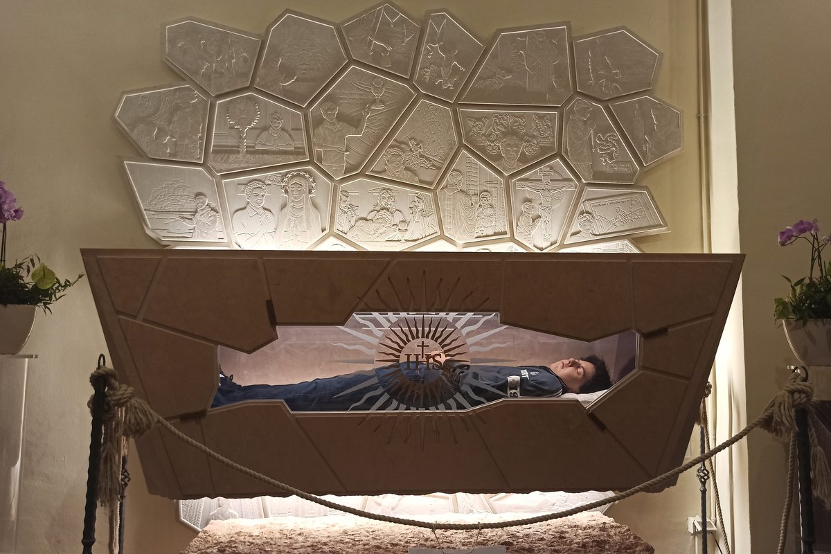 The glass-walled tomb of Carlo Acutis in the church of Santa Maria Maggiore in Assisi, showing his body dressed in jeans and sneakers beneath a radiant IHS emblem, with a mosaic depicting scenes from his life mounted on the wall above.