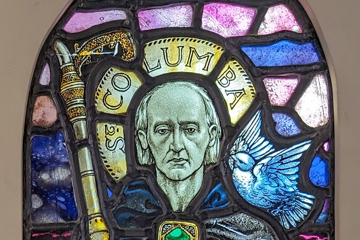 A stained-glass window depicting a tonsured monk saint holding a crosier and an illuminated Gospel book, with a dove beside his halo and the name Columba inscribed above.