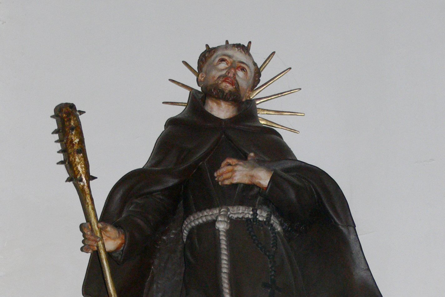 A polychrome statue of Saint Fidelis of Sigmaringen in a Capuchin habit, crowned with a martyr's radiant halo, gazing upward while holding a spiked club, a relic of his violent death.