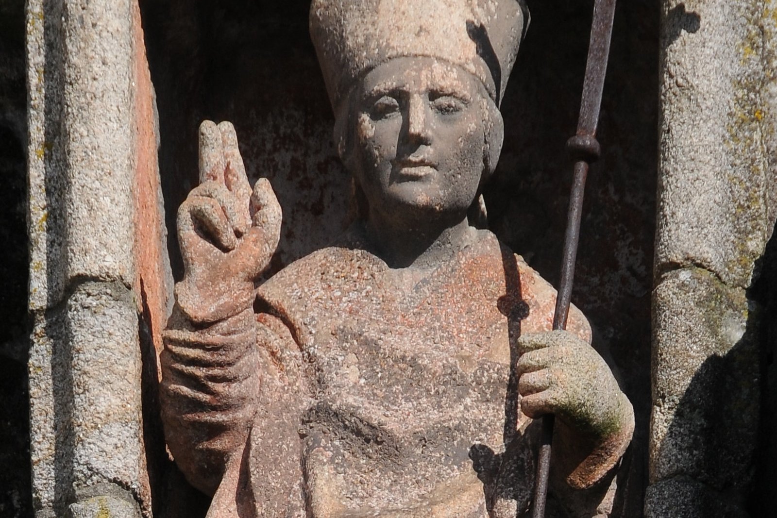 A bronze statue of a bearded bishop in flowing vestments, holding a crosier, set against a stone church wall.