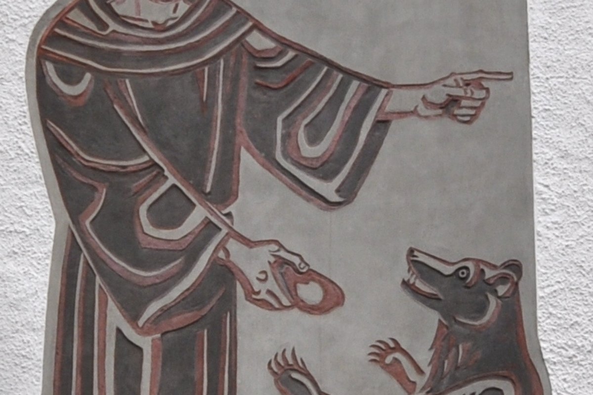 A relief carving on a wall in St. Gallen, Switzerland, inscribed "ST. GALLUS A.D. 612," showing the saint in a monk's habit pointing toward a bear that is standing on its hind legs beside a fire.