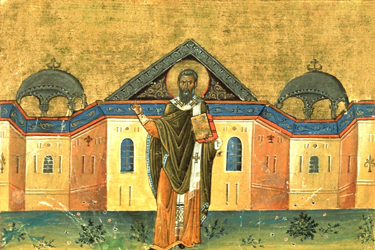A Byzantine manuscript miniature of a bearded bishop saint with a gold halo, wearing dark-green and orange vestments, blessing with one hand and holding a jeweled Gospel book, standing before a domed building.