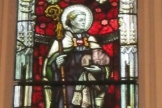 A stained glass window depicting a robed monastic saint holding a book and staff, in a church interior setting.