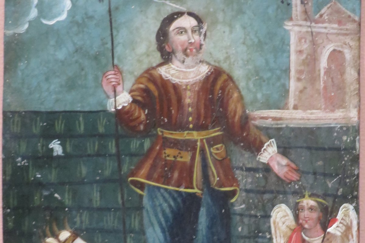 A folk-art retablo painting of a farmer in 17th-century dress standing between farmland and a small church, with a haloed angel and a yoked ox beside him.