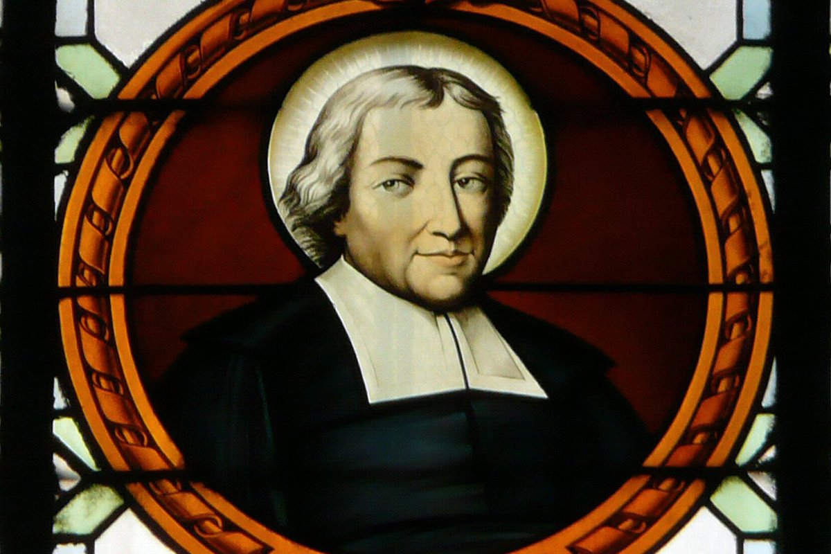 A stained-glass roundel portrait of a haloed priest with pale hair, wearing a black cassock and white clerical collar, labeled "St. J-B de la Salle."