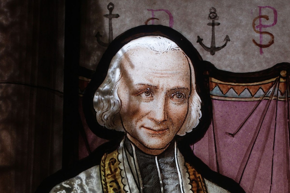 A stained glass window depicting an elderly priest with white hair, wearing ornate vestments trimmed in gold.