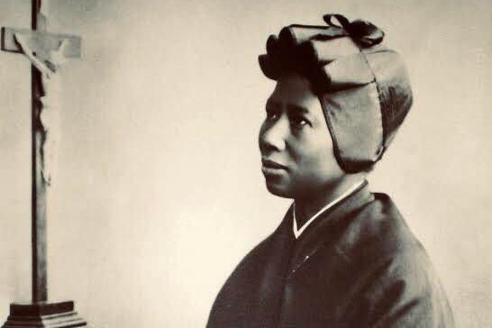 Sepia photograph of Saint Josephine Bakhita in her Canossian religious habit, kneeling in prayer before a crucifix, circa 1910