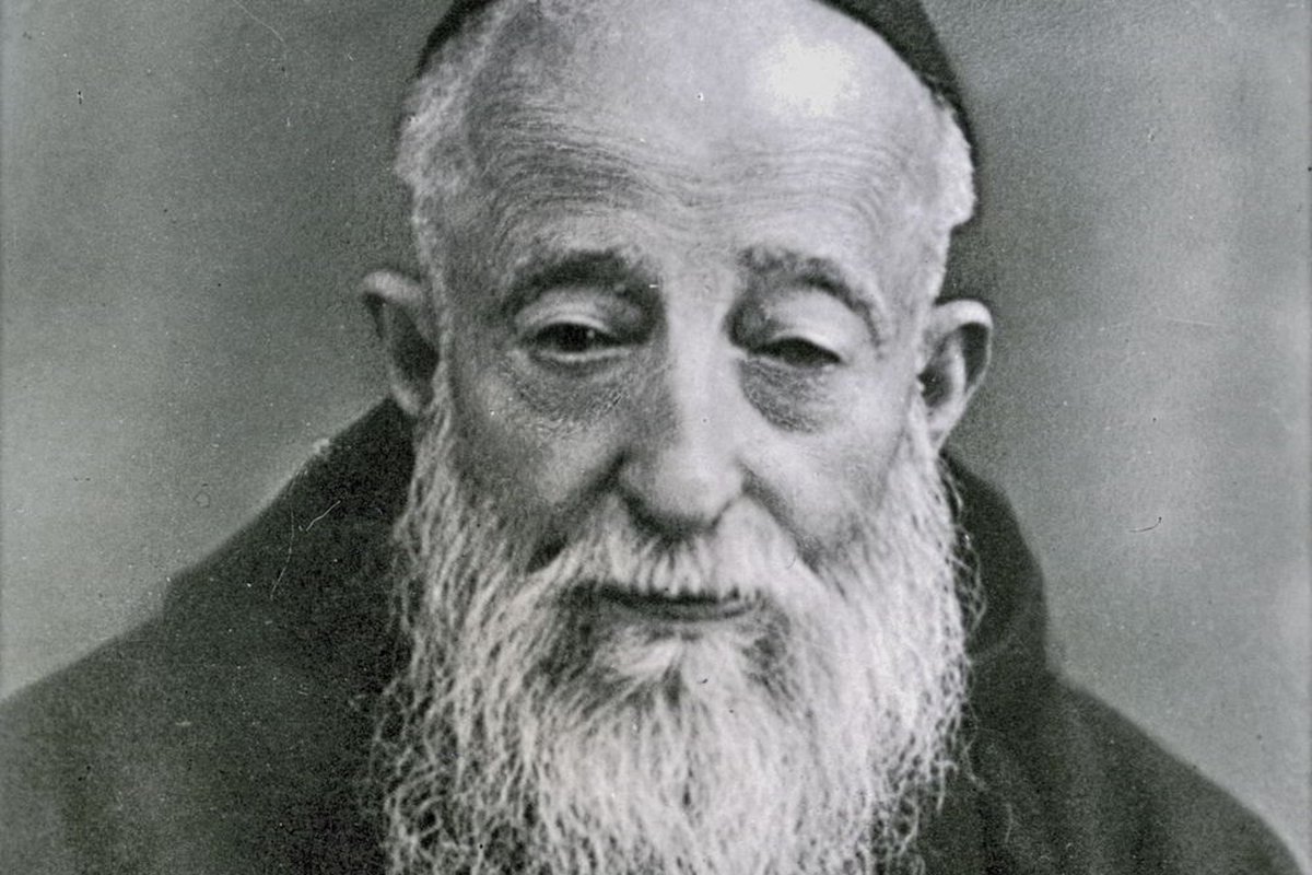 A black-and-white photographic portrait of an elderly Capuchin friar with a long white beard, wearing a dark habit and skullcap, looking downward.