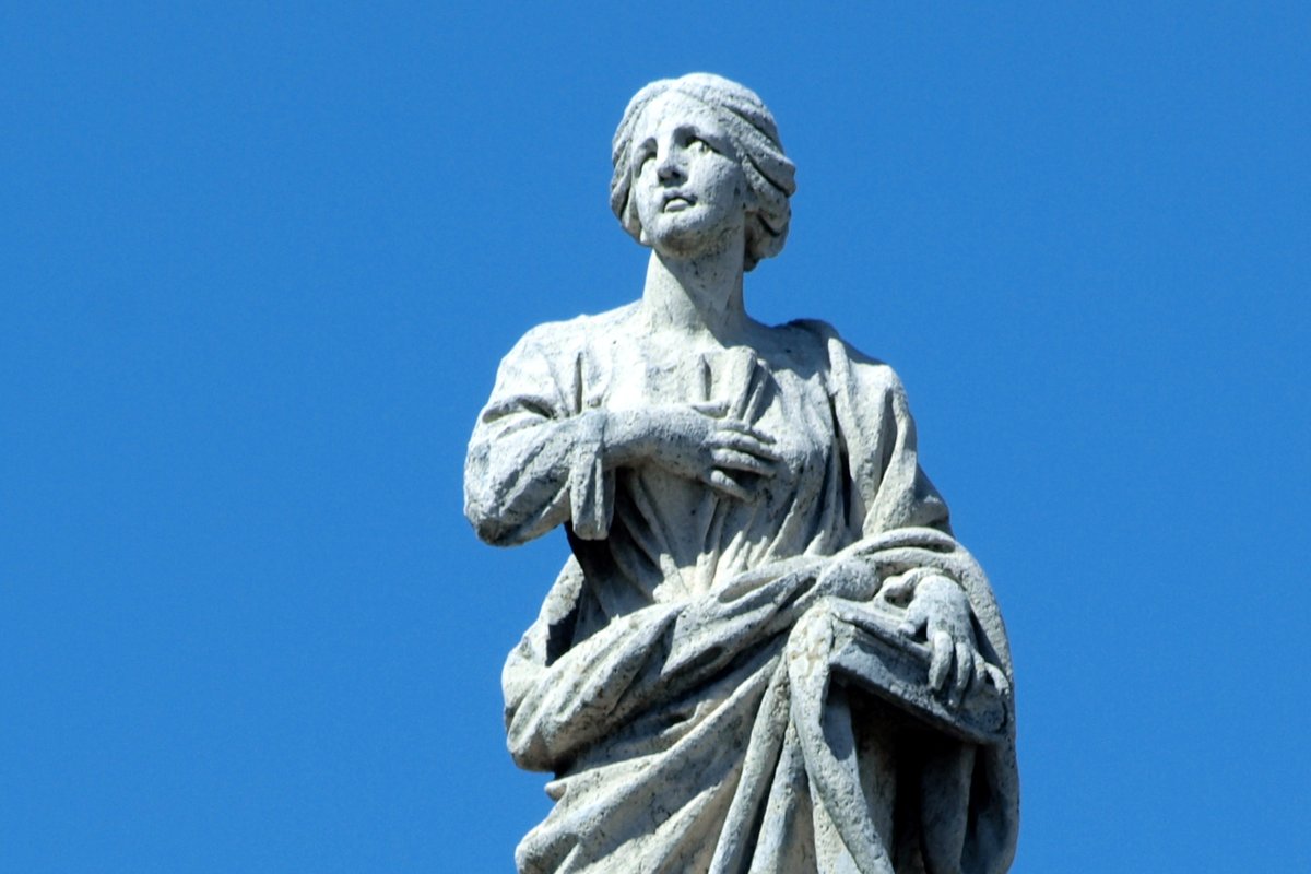 A weathered stone statue of a haloed young woman with her hand pressed to her chest, gazing upward, on the colonnade of Saint Peter's Square in Rome.