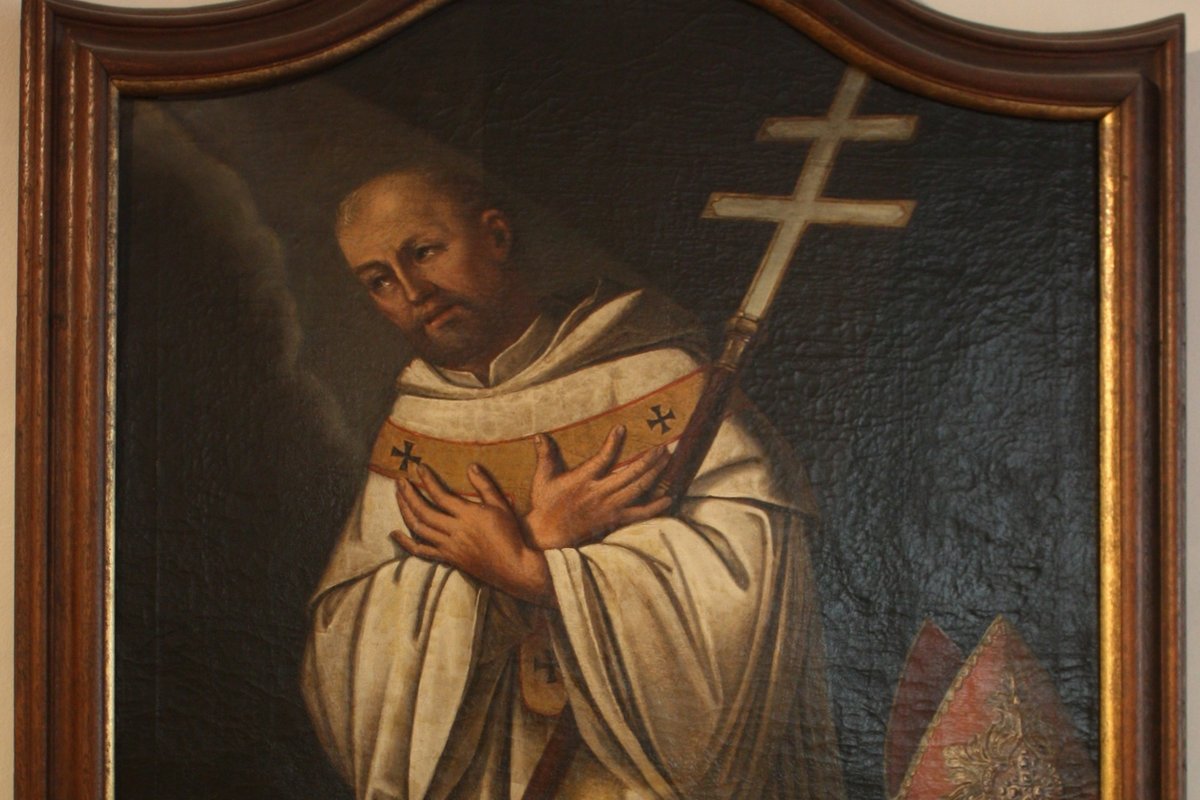 A dark oil portrait of a bald, bearded monk-bishop in white Cistercian habit, clutching a double-barred archiepiscopal cross to his chest, with a mitre resting beside him.