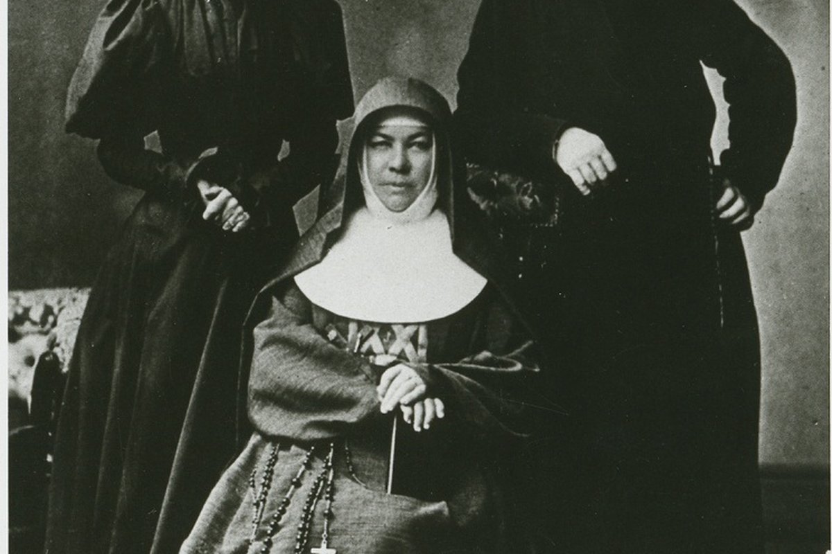 An 1890 photograph of Sister Mary MacKillop seated in her brown Josephite habit, holding a rosary and cross, with two companions standing beside her.
