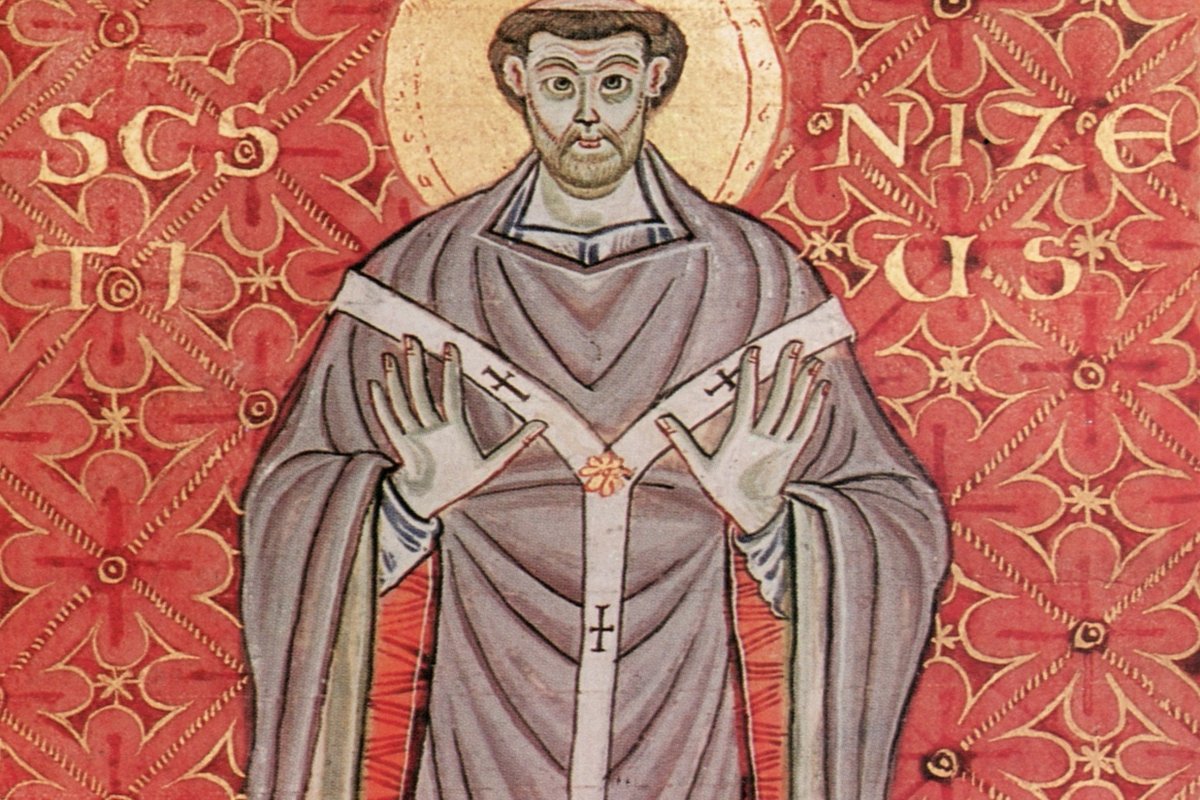 A 10th-century manuscript illumination of a haloed bishop in purple vestments, hands raised, flanked by gold lettering reading "SCS NIZETIUS" against a red patterned background.