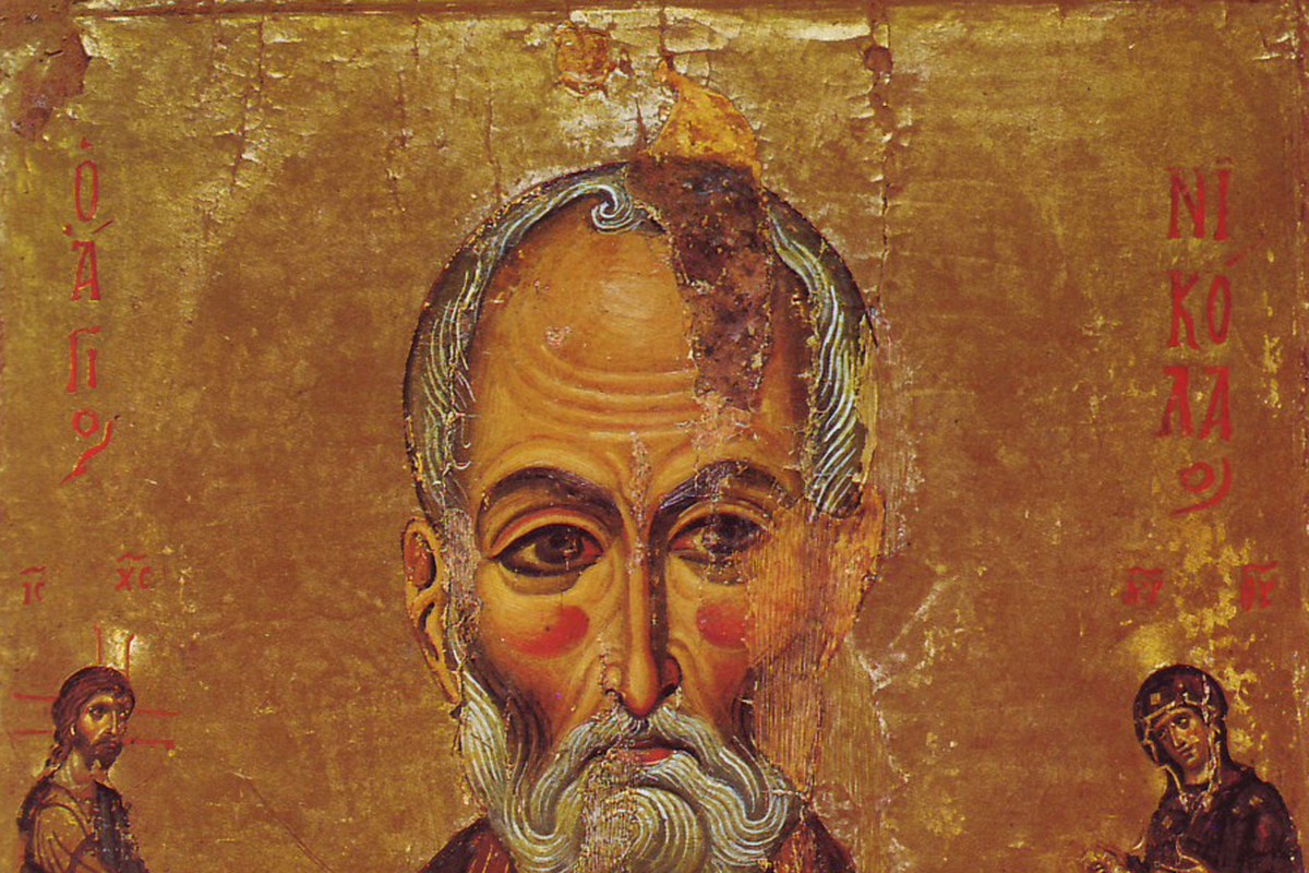 A traditional Byzantine-style icon of an elderly bishop in ornate robes, holding a book and raising a hand in blessing.