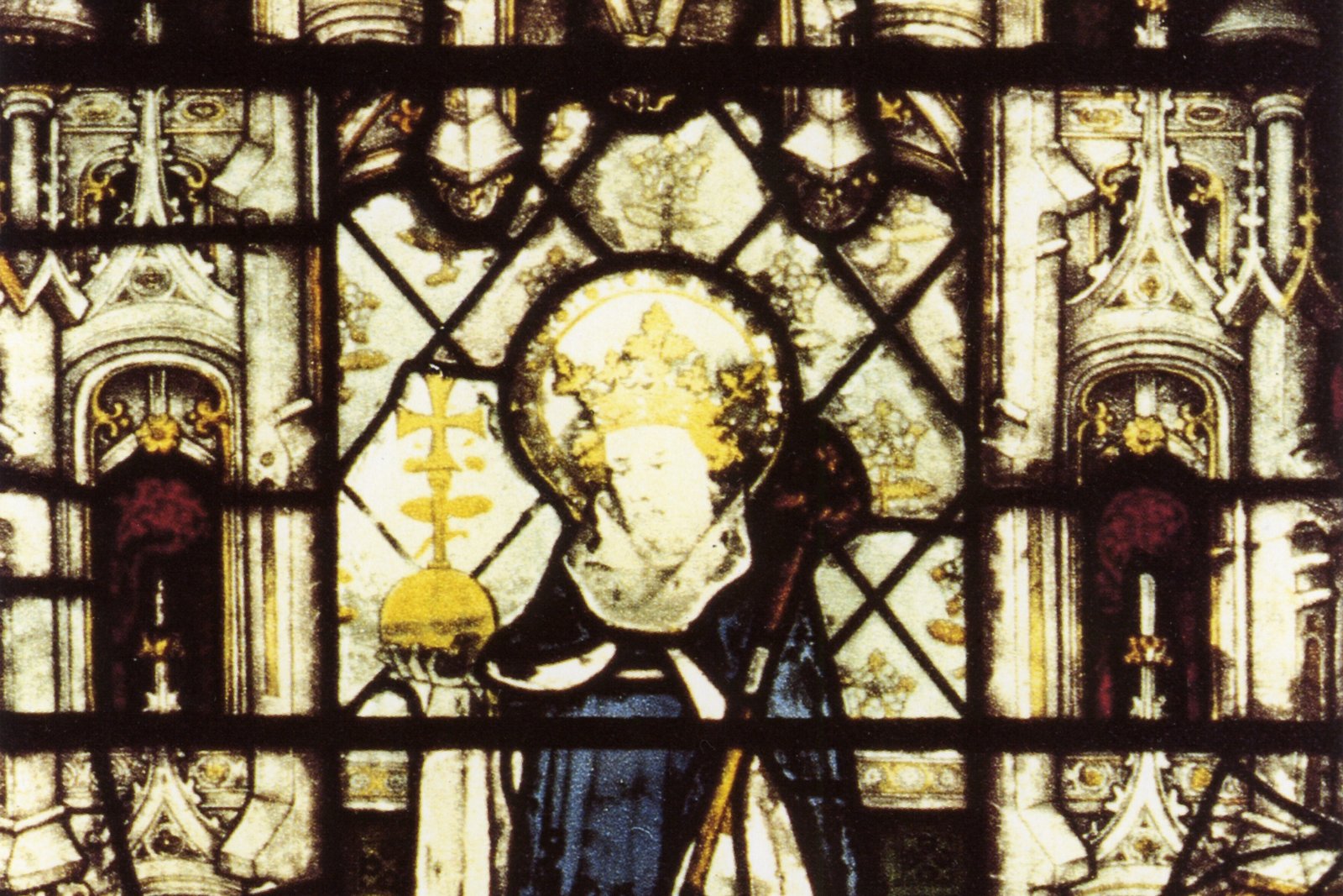 A stained glass window depicting a crowned, haloed bishop or king figure in blue and gold vestments, holding a cross-topped staff, from a Gothic chapel window at All Souls College, Oxford.