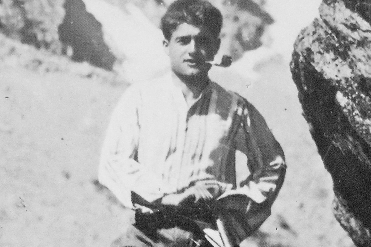 A black-and-white photograph of a young man in mountaineering clothes, smoking a pipe, standing on a rocky slope with an ice axe.