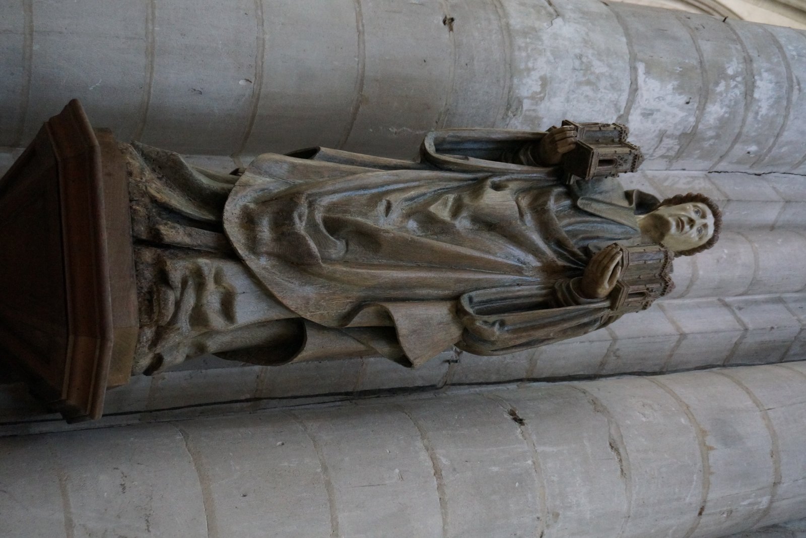 A painted wooden statue of a medieval abbot in monastic robes, holding a crosier, mounted against a stone wall.