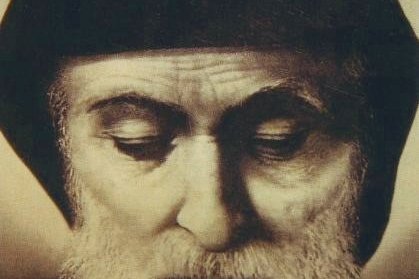 A close-up sepia-toned photograph of Saint Sharbel Makhluf, a bearded Maronite monk in a dark hood, eyes lowered in prayer.