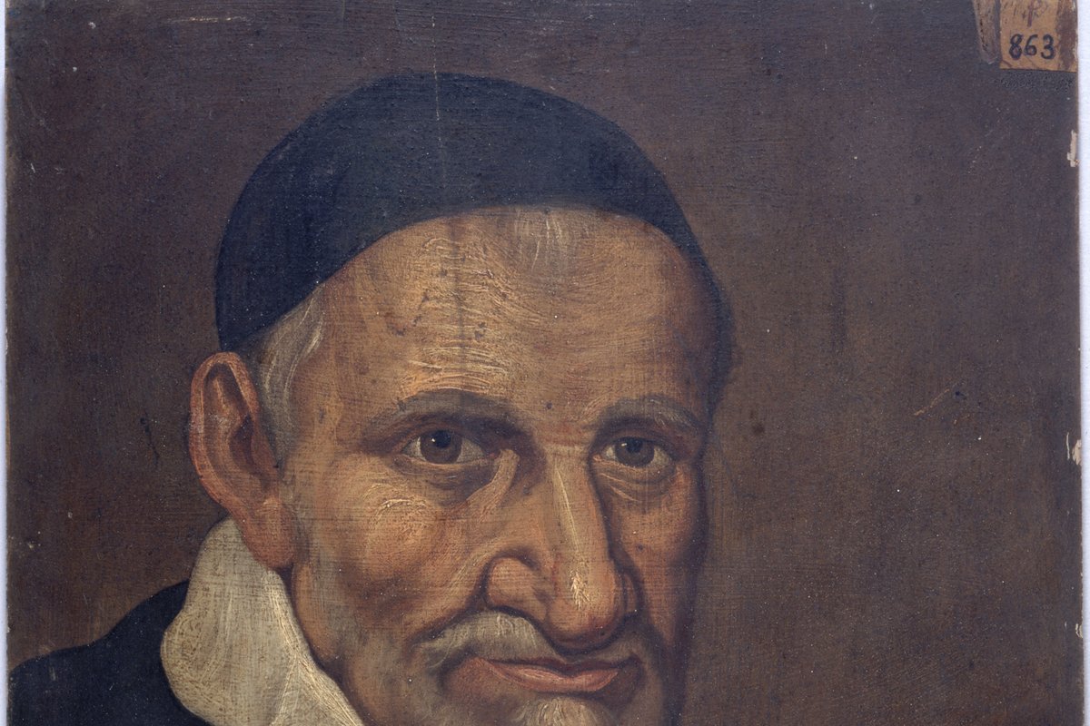A close portrait of an elderly priest with a lined, weathered face, white beard and moustache, wearing a black cap and a plain white clerical collar.