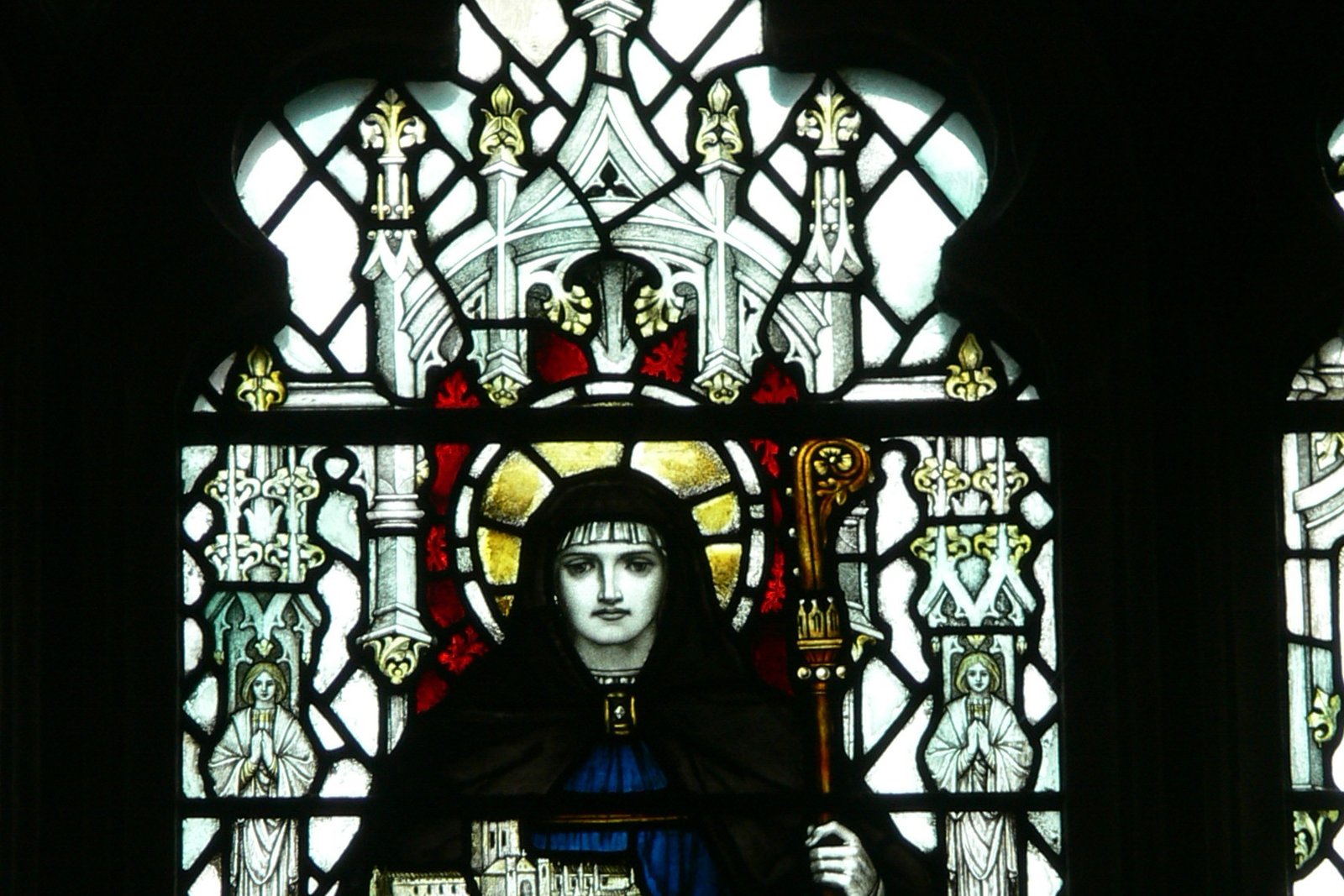 A stained-glass window depicting a haloed abbess-saint in dark robes holding a crozier, with a small walled city at her feet.