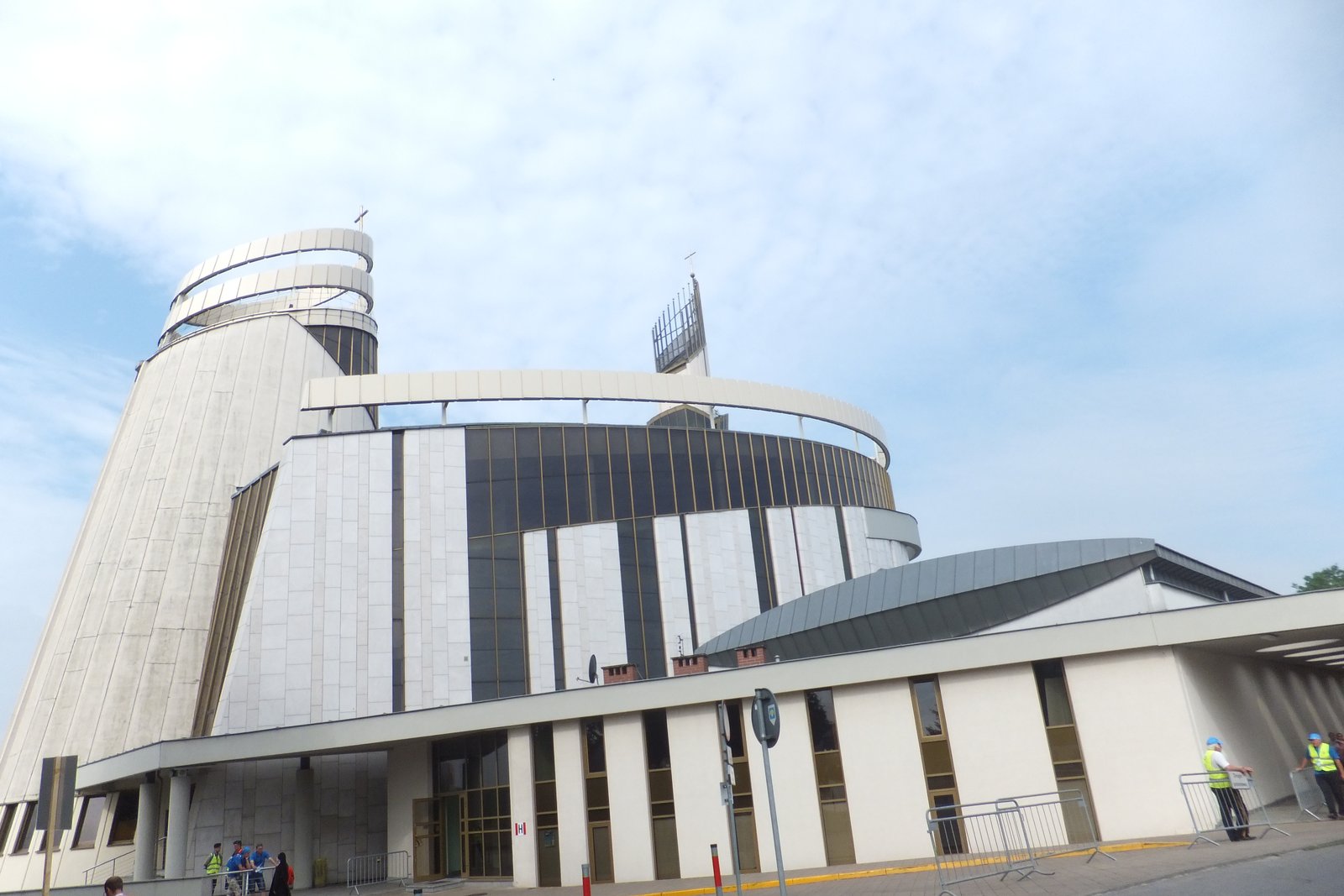 The modern curved white facade and glass tower of the Divine Mercy Sanctuary in Łagiewniki, Kraków, against a partly cloudy sky.