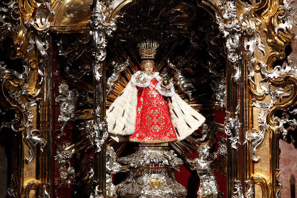 The small wax-coated wooden statue of the Infant Jesus of Prague, dressed in an ornate robe and crown, displayed at the Church of Our Lady Victorious