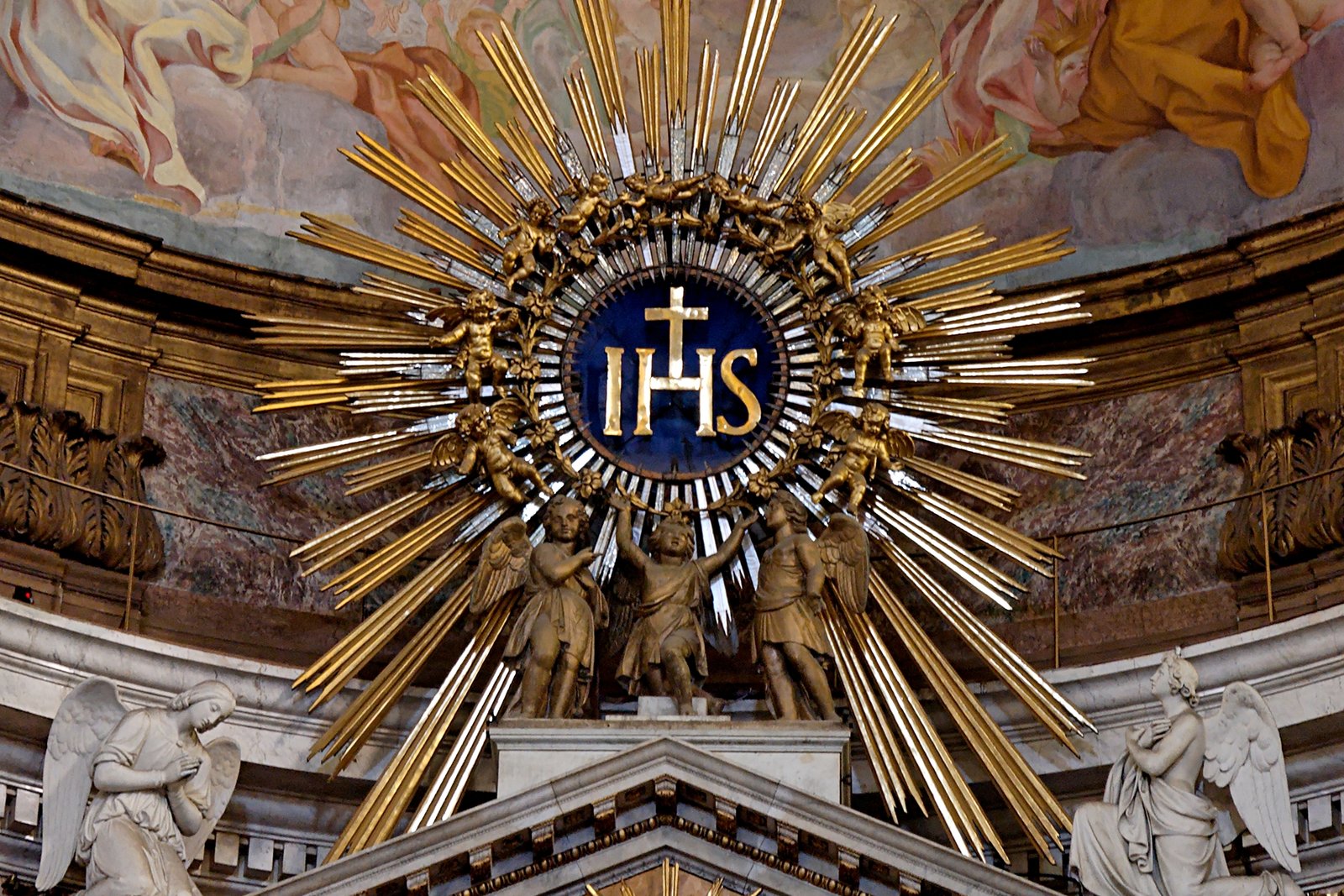 The golden IHS monogram of the Holy Name of Jesus surrounded by a sunburst and gilded cherubs above the high altar of the Church of the Gesù in Rome.