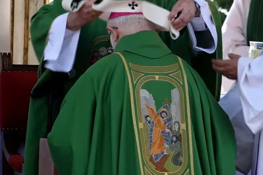 An archbishop kneeling as a white woolen pallium, marked with dark crosses, is placed around his shoulders during a liturgical ceremony