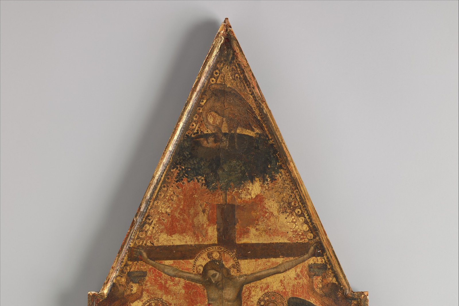 A gold-ground medieval altarpiece panel showing a pelican perched above a crucifixion scene, at the peak of the triangular panel.