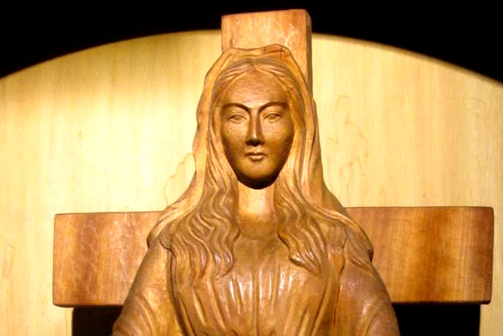Close-up of the wooden statue of Our Lady of Akita, carved with the face of a Japanese woman, hands open at her sides, standing before a wooden cross