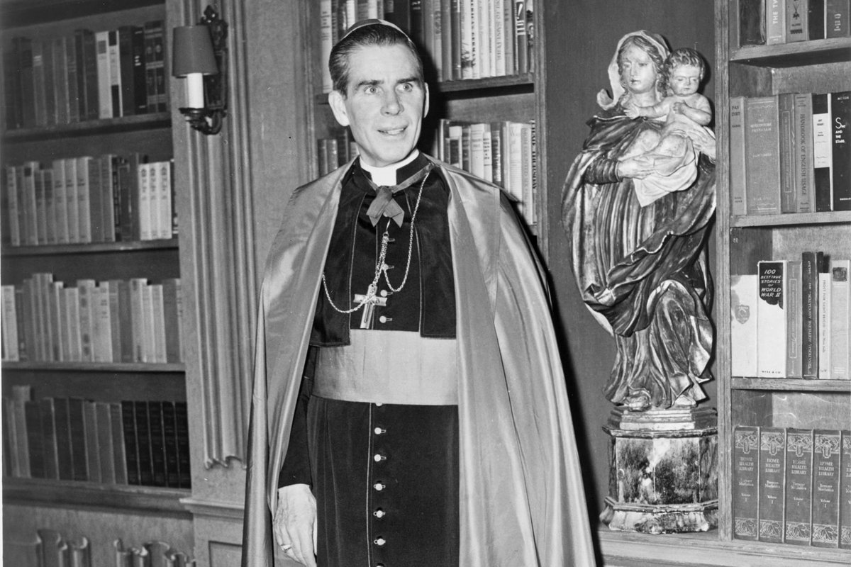A bishop in a black cassock and cape stands in a wood-paneled library beside a carved statue of the Madonna and Child, bookshelves lining the walls behind him.